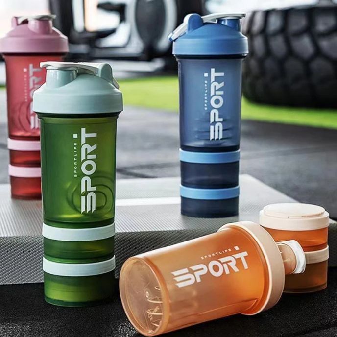 quatre shaker proteine de différentes couleurs (vert, bleu, orange,rose) alignés sur un tapis de sport à la salle de sport
