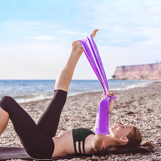  Femme allongée sur une plage faisant des exercices de jambes avec un elastique musculation violet