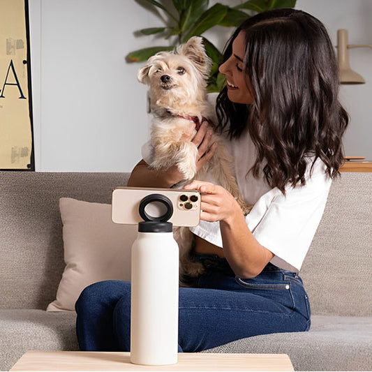 femme au salon avec gourde sport blanche et un telephone attaché à la gourde par aimant en face d elle et elle tient son chien entre les mains