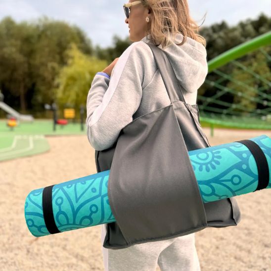 femme porte sac yoga gris sur son épaule avec tapis de yoga dans les manches latérales du sac et en tenue de sport dans un parc