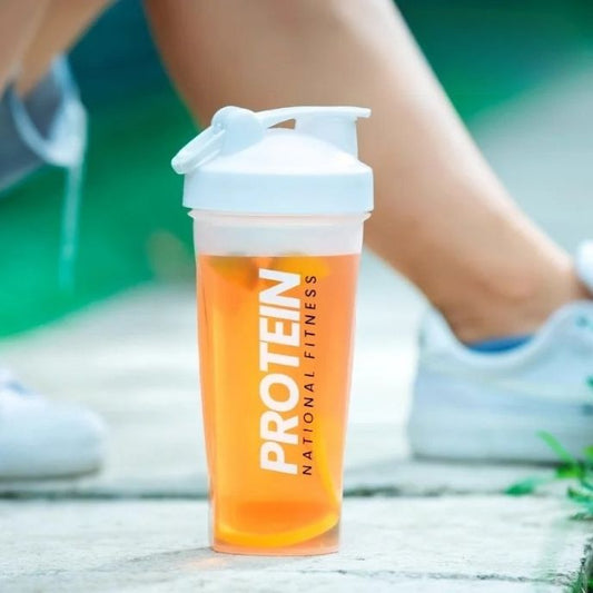 Shaker blanc avec boisson protéinée orange posé à côté d un genou en tenue de sport sur terrain vert