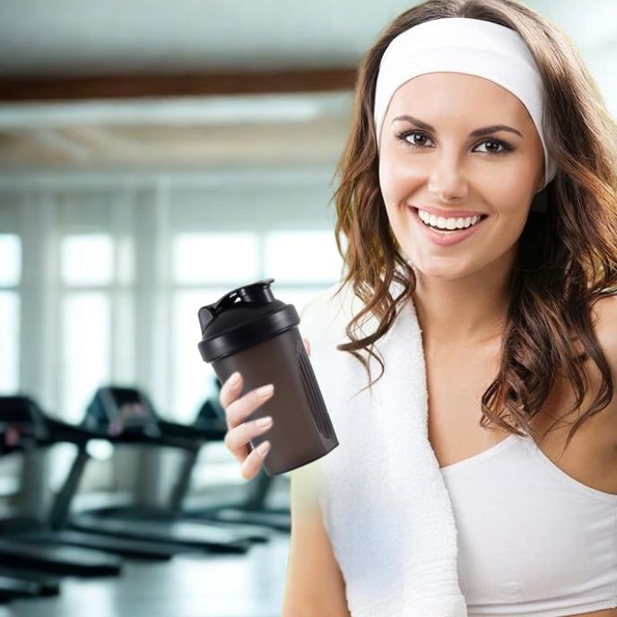 jeune femme en salle de sport tenant un shaker proteine noir et portant un bandeau blanc