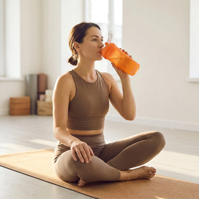 femme boit de shaker orange assise sur tapis de yoga